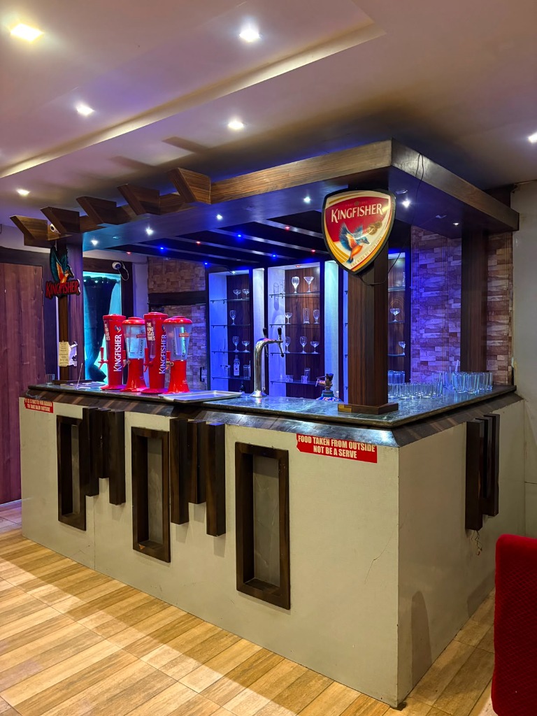 Modern bar counter with wooden pergola canopy, illuminated Kingfisher neon, draft beer dispenser towers, and back-lit cocktail glassware at Shah Junction Villa