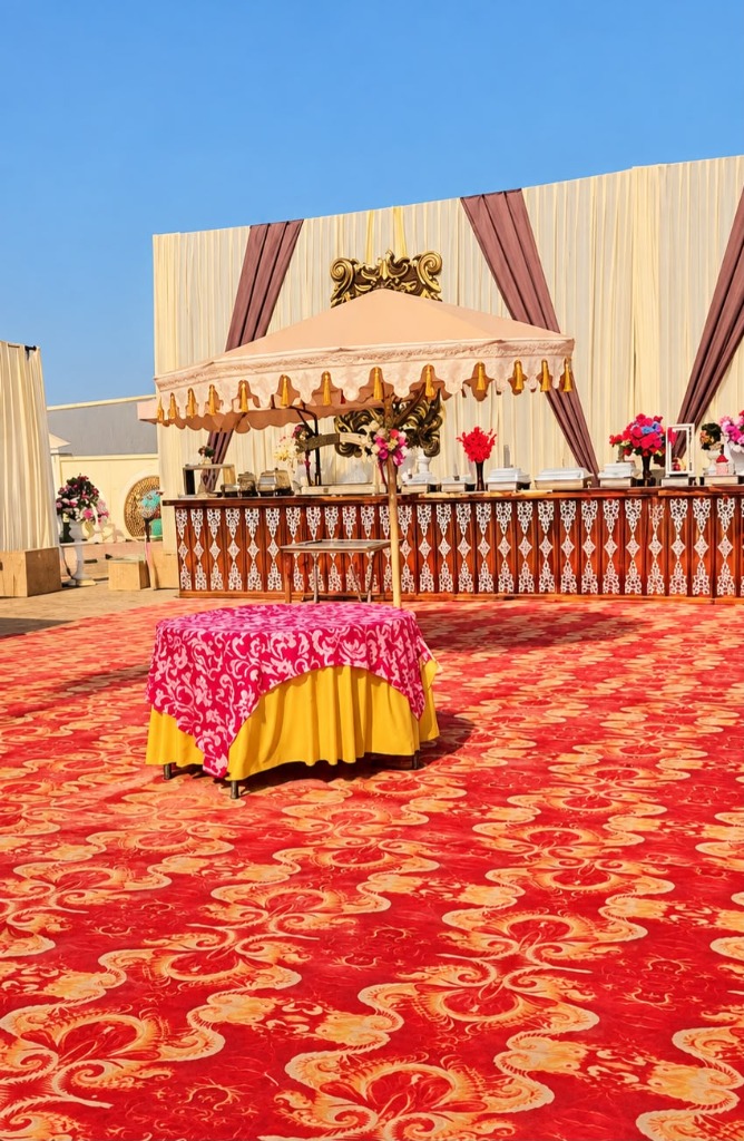 Outdoor celebration lawn catering counter with traditional wood carving, scalloped umbrella canopy, and red carpet runner under blue Punjab skies
